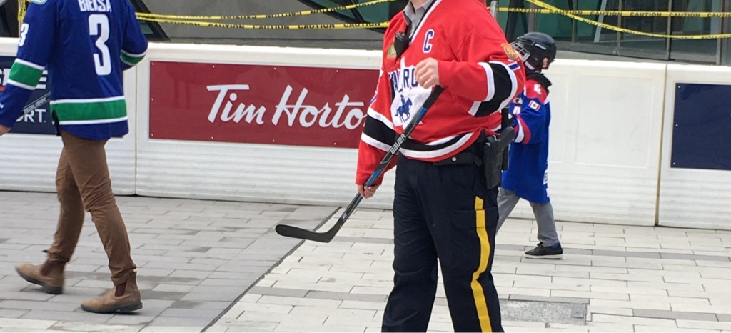 A RCMP playing road hockey at Hometown Hockey in Surrey, BC. He has a RCMP jersey on, but is also in dress pants and has a gun in his holster.