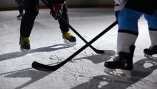 Frame of lower half of two hockey players with sticks on ice, facing off. Player one in mostly black jersey and player two in mostly white jersey.
