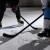 Frame of lower half of two hockey players with sticks on ice, facing off. Player one in mostly black jersey and player two in mostly white jersey.