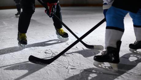 Frame of lower half of two hockey players with sticks on ice, facing off. Player one in mostly black jersey and player two in mostly white jersey.